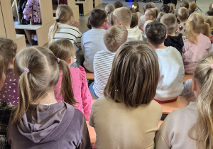 Dzieci siedzą na ławkach i patrzą na kobietę, która trzyma fagot.