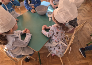 Dzieci siedzą przy stole w czapkach kucharskich i fartuszkach.