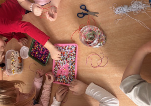 Dzieci przy stoliku nawlekają koraliki , tworząc bransoletki.