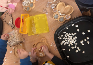 Dzieci przy stoliku tworzą wysypywanki z różnych materiałów , m.in fasoli, koralików.