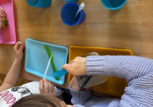 Dzieci przy stoliku, na nim kolorowe tace, polewają pipetą zamrożone dinozaury.