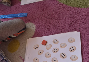 Dzieci układają dary zabawy Froebla na karcie pracy, na której znajdują się jajka oznaczone różnymi cyframi oraz różną ilością kropek.