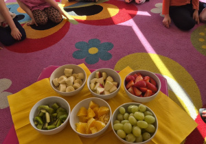 Dzieci siedzą w kole. Na środku stolik, na którym poukładane owoce w miseczkach.