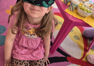 Dziewczynka siedzi na dywanie z zasłoniętymi oczami.