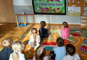 Dzieci siedzą na dywanie i oglądają na monitorze multimedialnym film edukacyjny o piramidzie zdrowego żywienia.