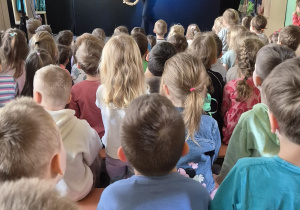 Dzieci siedzą na ławeczkach i oglądają przedstawienie teatralne