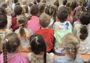 Dzieci siedzą na ławeczkach i oglądają spotkanie baletowe.