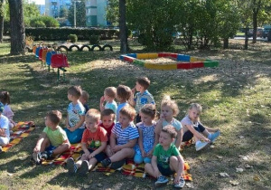 - Dzieci siedzą w ogródku przedszkolnym na kocu, rozłożonym na trawie
