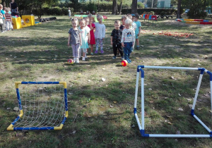 - Dzieci ustawione w dwóch rzędach przygotowują się do kopnięcia piłki do bramki