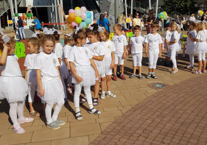 Grupa dzieci w białych strojach stoi na chodniku przed Centrum Handlowym Port Łódź