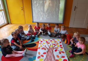 Dzieci siedzą w kole i prezentują wykonaną przez siebie pracę przedstawiającą drzewo.