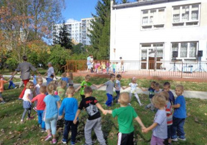 Ogród przedszkolny. Na pierwszym planie grupa dzieci w kole porusza się w jednym kierunku trzymając za ręce. W oddali druga grupa dzieci z nauczycielką. Na tarasie kolejna grupa dzieci z nauczycielką.