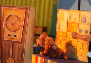 Marionetka przedstawiająca psa przed regałem z półkami.