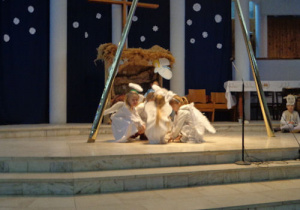 Na środku sceny kucają trzy dziewczynki w strojach aniołków.