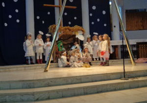 Józef z Maryją w szopce. Obok nich stoją dzieci w strojach aniołków. Przed nimi pastuszkowie.