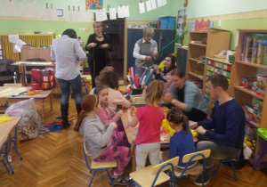 Dzieci i rodzice siedzą przy stole z bibułami. Trzy kobiety stoją wokół. Niektóre dzieci trzymają w dłoniach skrawki bibuły.
