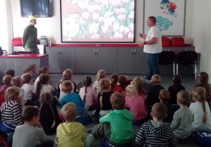 Dzieci siedzą na dywanie i patrzą w kierunku ekranu, na którym wyświetlony jest slajd z kwiatami. Obok stoi dwóch mężczyzn.