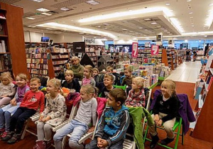 Dzieci siedzą na krzesełkach i patrzą w jednym kierunku. W tle regały z książkami.