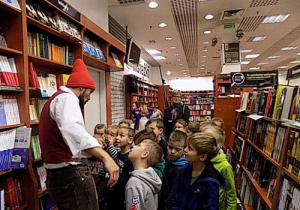 Dzieci stoją między regałami z książkami i przyglądają się mężczyźnie w czerwonej czapce.