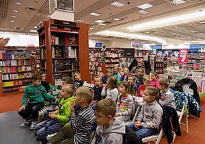 Dzieci siedzą na krzesełkach i patrzą w jednym kierunku. W tle regały z książkami.