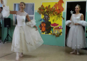 Dwie tancerki w białych, długich tiulowych sukienkach trzymają w rękach różdżki ze srebrną gwiazdką. Jedna z nich unosi różdżkę w górę.