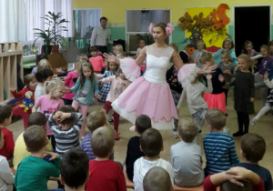 Tancerka w biało różowej sukience stoi na jednej nodze i przechyla lekko w lewo. Za kobietą stoją dziewczynki i naśladują jej ruch. Chłopcy na ławeczkach przyglądają się im.
