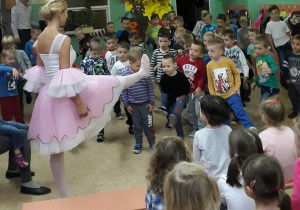 Tancerka w biało różowej sukience unosi wysoko przed siebie prawą nogę. Przed kobietą stoją chłopcy i naśladują jej ruch.