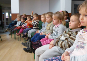 Dzieci siedzą na krzesłach ustawionych jedno obok drugiego. Jedno z dzieci podnosi rękę do góry.