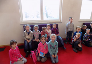 Dzieci w holu siedzą na kanapach i czerwonym dywanie.