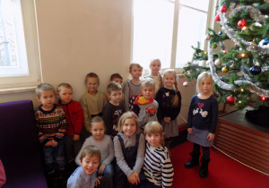 Dzieci w holu siedzą na kanapach i czerwonym dywanie. Obok dzieci świątecznie udekorowana choinka.