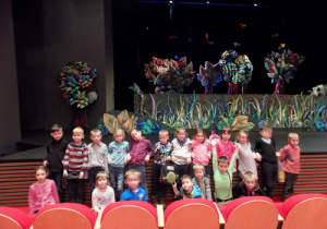 Grupa dzieci pozuje przed sceną. W tle kolorowa scenografia.