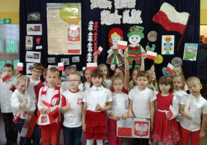 Dzieci stoją w holu przedszkolnym w strojach o barwach narodowych pod napisem „Piękna Nasza Polska Cała”
