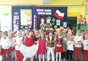 Duża grupa przedszkolaków w biało-czerwonych strojach stoi w holu pod napisem „ Piękna Nasza Polska Cała”