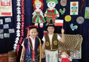 Dwóch chłopców w strojach ludowych stoi pod Tablicą Patriotyczną