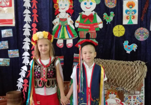 Dziewczynka i chłopiec w strojach ludowych stoją pod Tablicą Patriotyczną
