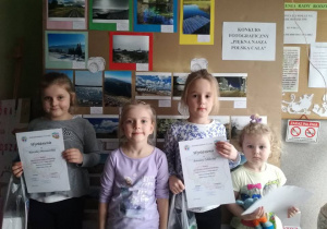 Dziewczynki z dyplomami za udział w konkursie stoją na tle zdjęć na wystawie