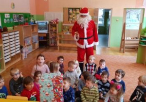 - Dzieci siedzą na dywanie z prezentem, a Mikołaj stoi