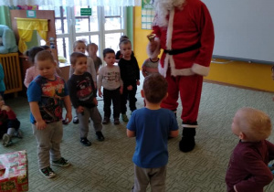 - Dzieci stoją z Mikołajem i prezentem na dywanie