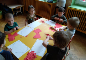 Dzieci przy stoliku wypełniają serca czerwonym papierem