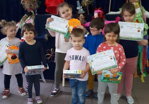- Dzieci stoją z dyplomami na tle wystawy Marzanny i napisu „ Już Wiosna”