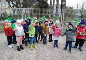 - Dzieci stoją przed ogrodzeniem przedszkolnym i machają gałązkami z zielonej bibułki