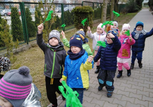 - Dzieci z zielonymi bibułkami idą przedszkolnym chodnikiem w kierunku furtki