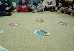 Dzieci siedzą w kole. Na dywanie w różnych miejscach położone są książki.