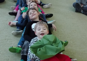 Dzieci siedzą w rzędzie, przechylają się do tyłu, trzymają się za ramiona. Pierwszy chłopiec trzyma w rzepkę zrobioną z poduszki.