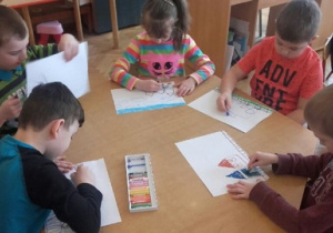 Pięcioro dzieci siedzi przy stoliku. Rysują pastelami. Kredki w pudełku znajdują się na środku stolika. Jeden chłopiec podnosi kartkę do góry.