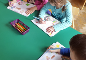 Troje dzieci rysuje przy stoliku, na którym leżą kredki ołówkowe we fioletowym koszyczku.