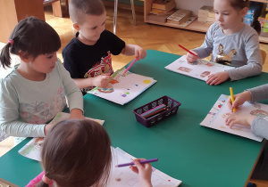 Czworo dzieci rysuje przy zielonym stoliku, na którym leżą kredki ołówkowe we fioletowym koszyczku. Widać rączki i rysunek czwartego dziecka.