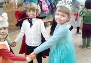 Dzieci w strojach wampira, księżniczki i lisa stoją w kole trzymając się za ręce.