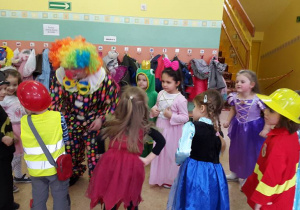 Dzieci w kolorowych strojach stoją w kole z nauczycielką przebraną za clowna.