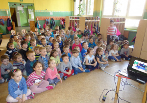 Dzieci siedzą z twarzami zwróconymi w stronę tablicy multimedialnej